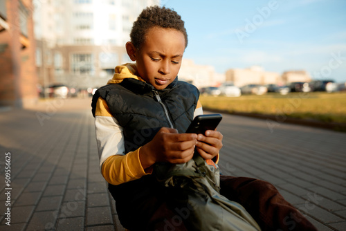 Cute adorable african american kid of 8-years-old playing game on smartphone sitting on sidewalk, losing track f time, returning home from school. Children, gadget and phone addiction