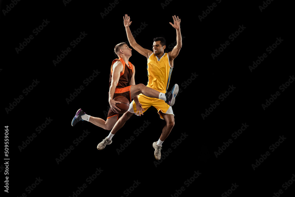 Dynamic portrait of two young men, professional basketball players in a jump, throwing ball into basket isolated over black studio background.