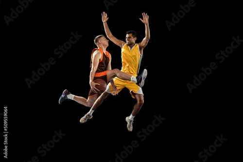 Dynamic portrait of two young men  professional basketball players in a jump  throwing ball into basket isolated over black studio background.