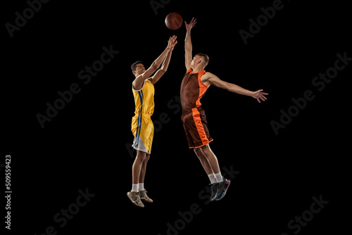 Dynamic portrait of two young men, professional basketball players in a jump, throwing ball into basket isolated over black studio background.