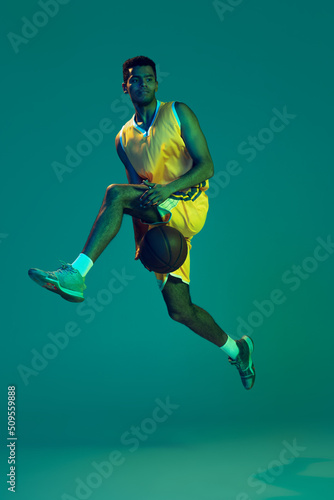 Portrait of young man, professional backetball player in motion, training isolated over blue background in neon light © Lustre
