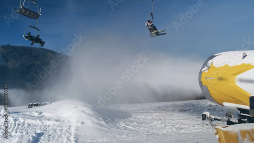 Snow cannon makes artificial snow on background of working ski lift with people, going to mountain slope in sunny day. Snow making system blows water in ski resort, backdrop blue sky