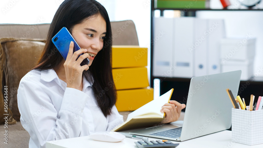 Work from home, Young asian woman talking mobile phone and holding notebook while working at home office, People and technology communication, e commerce, online business, e learning, education Work from home, Young asian woman talking mobile phone and holding notebook while working at home office, People and technology communication, e commerce, online business, e learning, education