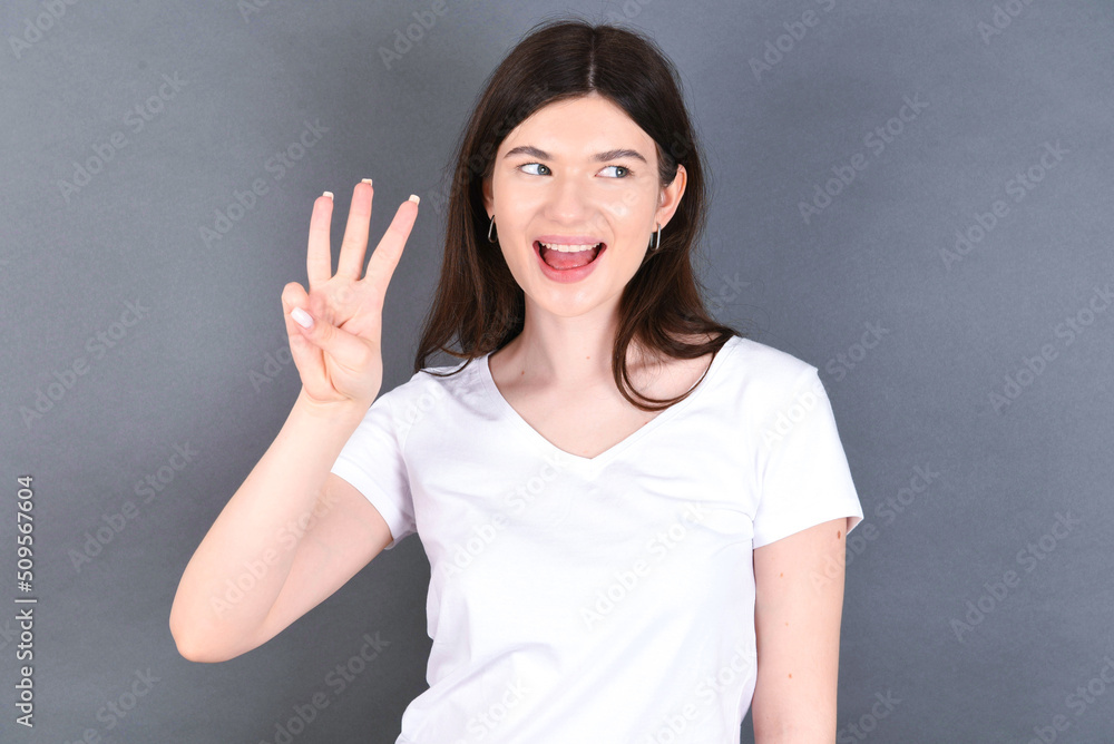 young beautiful Caucasian woman wearing white T-shirt over studio grey wall showing and pointing up with fingers number three while smiling confident and happy. young beautiful Caucasian woman wearing white T-shirt over studio grey wall showing and pointing up with fingers number three while smiling confident and happy.