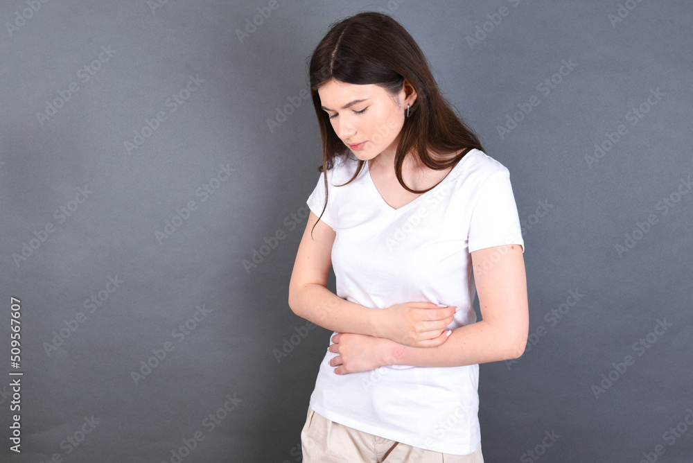 young beautiful Caucasian woman wearing white T-shirt over studio grey wall suffering from strong stomachache. young beautiful Caucasian woman wearing white T-shirt over studio grey wall suffering from strong stomachache.