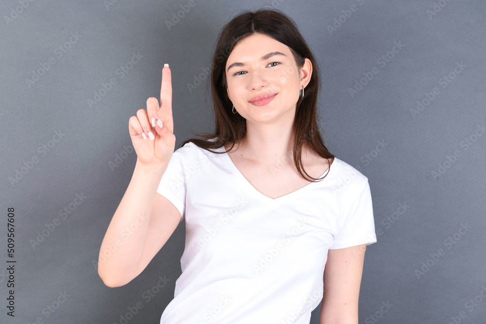 young beautiful Caucasian woman wearing white T-shirt over studio grey wall showing and pointing up with fingers number one while smiling confident and happy. young beautiful Caucasian woman wearing white T-shirt over studio grey wall showing and pointing up with fingers number one while smiling confident and happy.