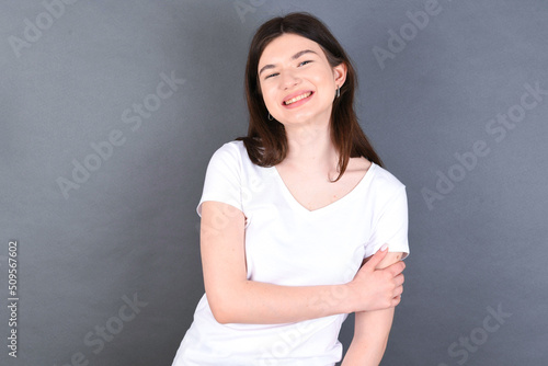 People, lifestyle, youth and happiness concept. Shy pretty young beautiful Caucasian woman wearing white T-shirt over studio grey wall, feeling happy hugging herself.