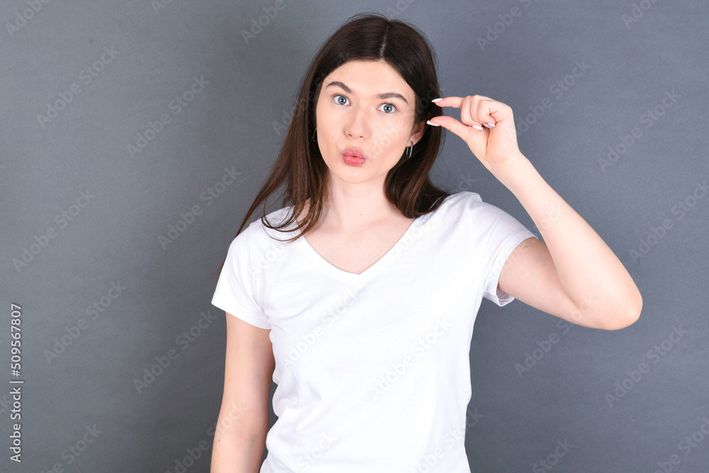 Shocked young beautiful Caucasian woman wearing white T-shirt over studio grey wall shows something little with hands, demonstrates size, opens mouth from surprise. Measurement concept. Shocked young beautiful Caucasian woman wearing white T-shirt over studio grey wall shows something little with hands, demonstrates size, opens mouth from surprise. Measurement concept.