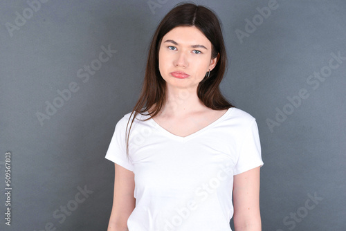 Gloomy, bored young beautiful Caucasian woman wearing white T-shirt over studio grey wall frowns face looking up, being upset with so much talking hands down, feels tired and wants to leave.