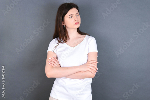 Image of upset young beautiful Caucasian woman wearing white T-shirt over studio grey wall with arms crossed. Looking with disappointed expression aside after listening to bad news.