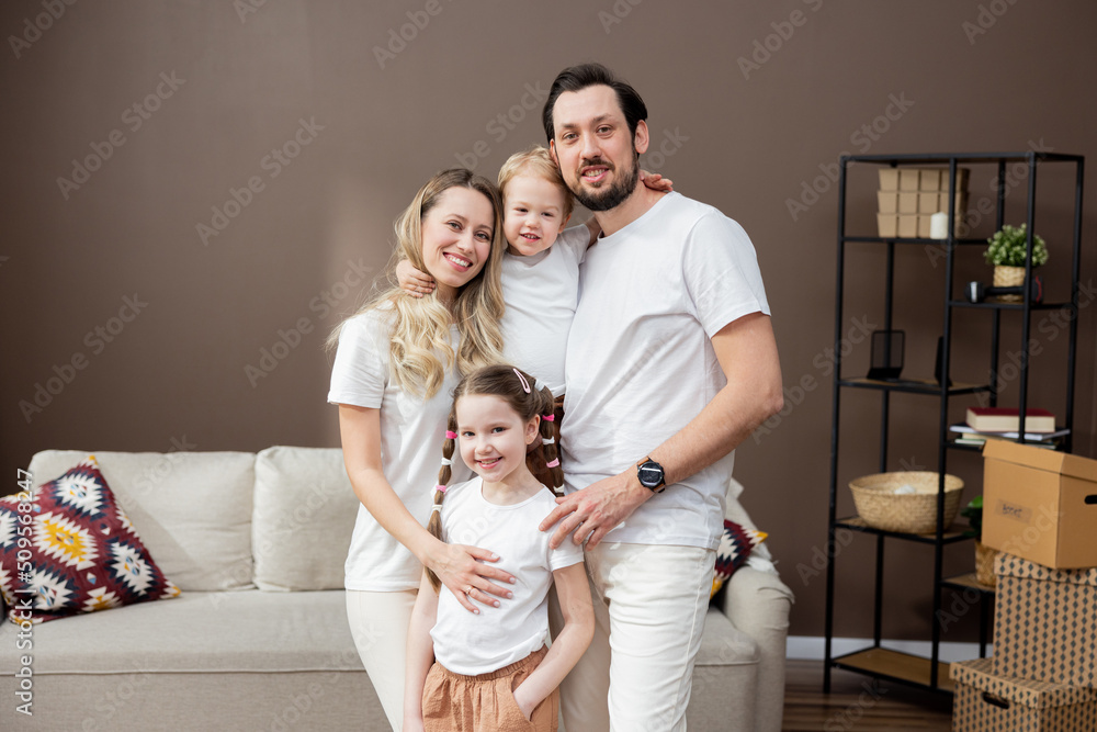 Portrait of happy young Caucasian family with kids standing in new home, holding cardboard boxes, smiling and looking at camera. Real estate, buying property, moving house or apartment concept.