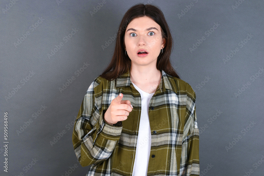 Shocked young beautiful Caucasian woman wearing plaid shirt over studio grey wall points at you with stunned expression Shocked young beautiful Caucasian woman wearing plaid shirt over studio grey wall points at you with stunned expression