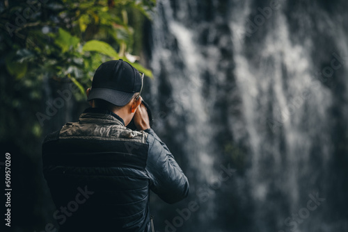 Close-up of back side photographer in casual style wear jacket and hat is using modern camera photography with blurred waterfall. 