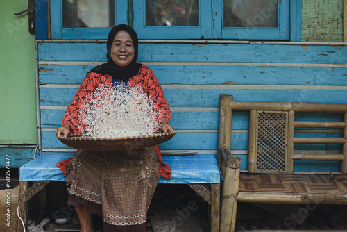 Bilde på lerret Asian local woman is sitting and sifting rice with bamboo colander against traditional wooden house