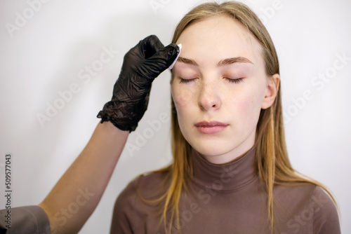 A young woman in a beauty salon uses the services of an eyebrow master. Eyebrow master in black gloves wipes eyebrows with lotion before coloring. Modern beauty care in a beauty salon