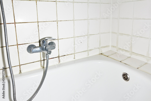 Spring cleaning concept: filthy shower armature in bath tub with black mold growing on calcifications on the tile grouting in an unclean old bathroom.