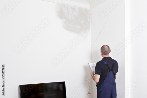 A janitor or custodian inspecting a big water stain and various cracks in wall and ceiling of the living room of an old building.
