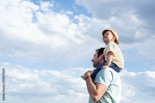 Side view of a smile little girl, looking up and sitting on her father's shoulders on a background of blue sky with clouds.