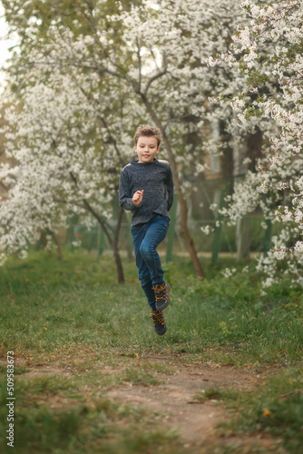 Cute boy in grey sweater and jeans is jumping in blooming cherry garden. Cute boy outdoor in the spring.