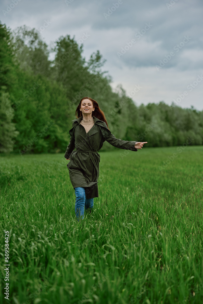 a beautiful woman in a long raincoat runs across a field in high grass in spring in cloudy weather