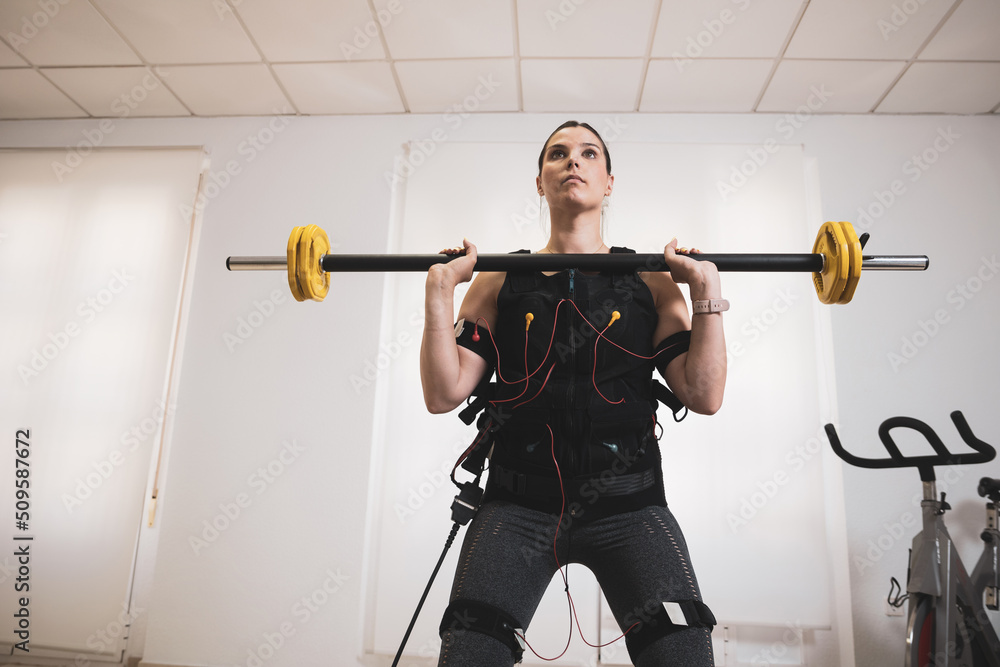 A beautiful young woman lifting weights while wearing an electrostimulation vest. A beautiful young woman lifting weights while wearing an electrostimulation vest.