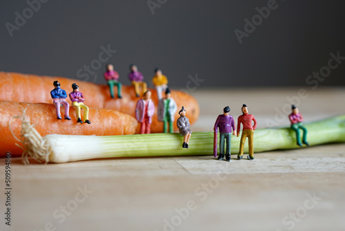 Miniature figurine character: people sitting on carrots and spring onions, and some standing on wooden floor, concept of fresh organic vegetables for good health in the market, selected focus