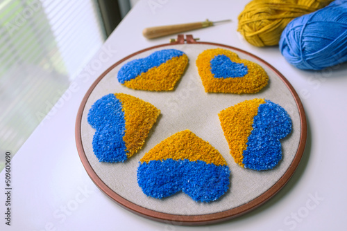Side view of fluffy tufted hearts on grey fabric in a hoop, punch needle and two balls of yarn in blue and yellow colors.