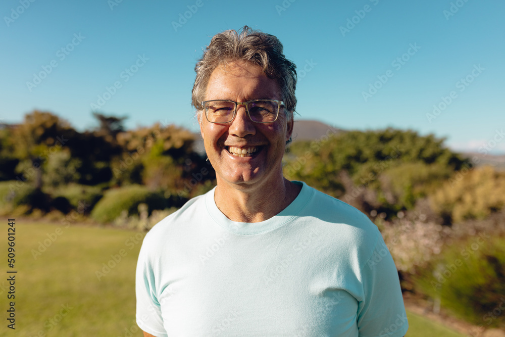 Portrait of cheerful caucasian senior man wearing eyeglasses standing against plants and sky in yard