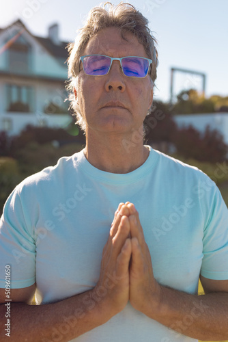 Caucasian senior man wearing eyeglasses with eyes closed meditating in prayer position in yard