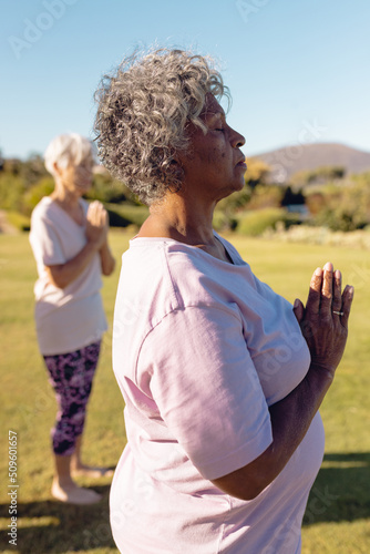 Multiracial senior women meditating in prayer position in yard against clear sky at retirement home