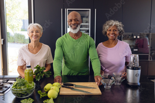 Portrait of smiling multiracial senior friends making apple and leaf vegetable smoothie in kitchen