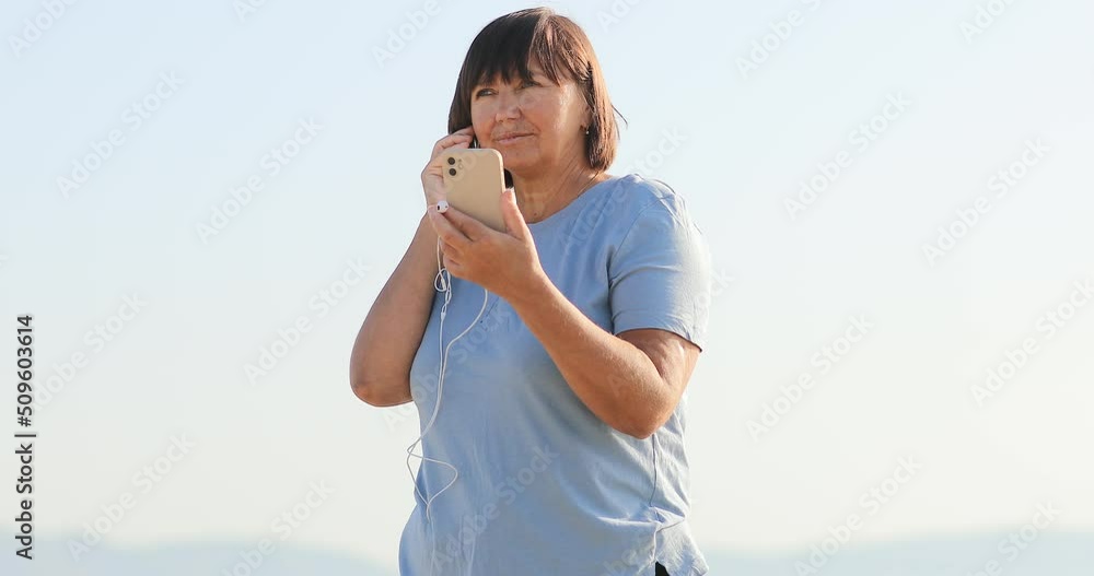 smiling middle aged woman talking on video call using smartphone with headphones on the beach. female 50 years remote has conversation with family on vacation. smiling middle aged woman talking on video call using smartphone with headphones on the beach. female 50 years remote has conversation with family on vacation.