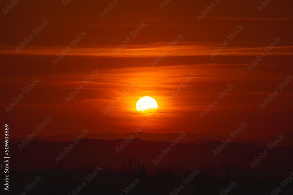 View of an orange sunset with the sun in the center above the colored clouds of orange and yellowish tones, in the lower part is the city
