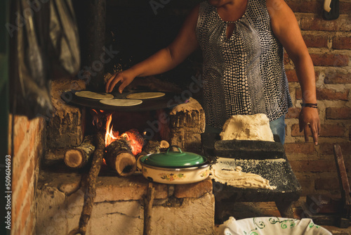 Mujer mexicana torteando maza de maíz en un metate y una estufa de leña para hacer tortillas caseras 