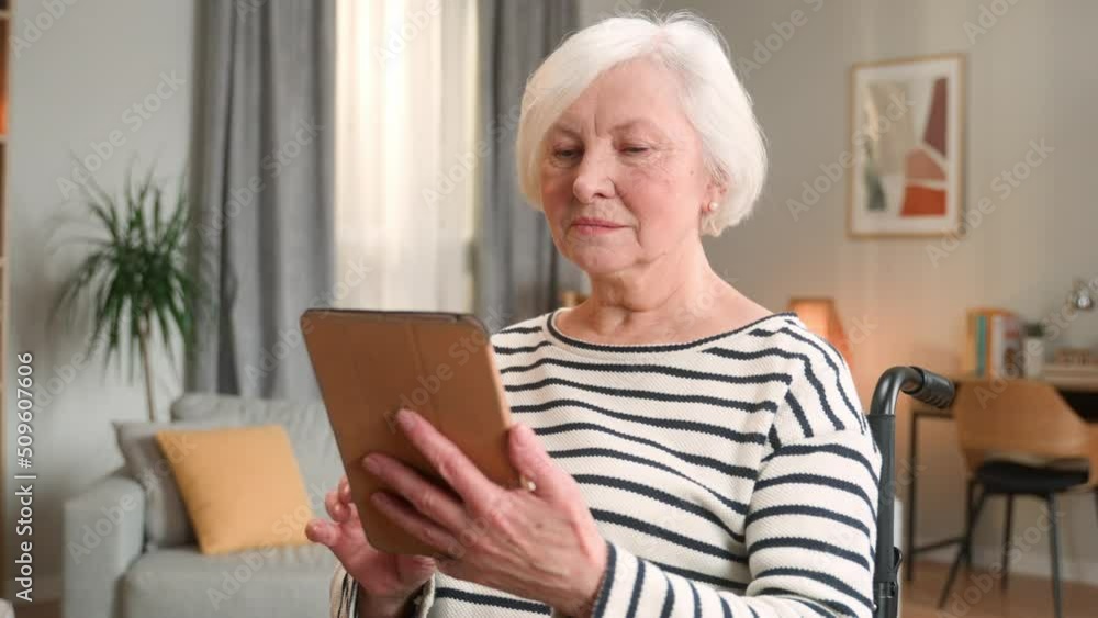 Close up of pretty senior woman in wheelchair in living room alone looking at tablet scrolling and tapping on gadget. Elderly happy lady using device surfing internet indoors. Disabled person concept Close up of pretty senior woman in wheelchair in living room alone looking at tablet scrolling and tapping on gadget. Elderly happy lady using device surfing internet indoors. Disabled person concept