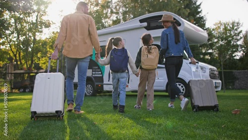 Rear view of young family with suitcases going to caravan outdoors at park.