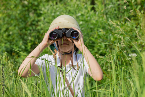 Preteen girl wearing pith helmet hiding in the grass looking through binoculars observing summer nature. Discovery and adventures concept photo