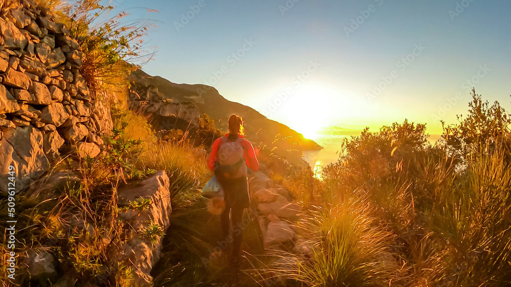 Active woman with backpack watching sunrise from hiking trail Path of Gods between Positano and Praiano, Amalfi Coast, Campania, Italy, Europe. Grass blowing in wind at golden hour, Mediterranean Sea