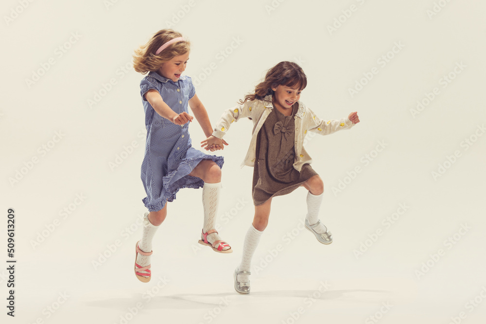 Portrait of two cheerful happy girls, children in beautiful dresses playing together, jumping isolated over grey studio background