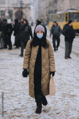 A young girl in a protective medical blue mask, in a brown down jacket, hat and hood, walks along a winter street and carries a package in her hand. Covid-19 coronavirus pandemic concept.Lviv, Ukraine © shchus