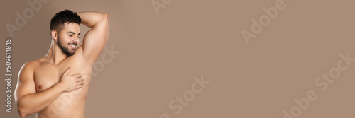 Young man showing armpit after applying deodorant on brown background, space for text. Banner design