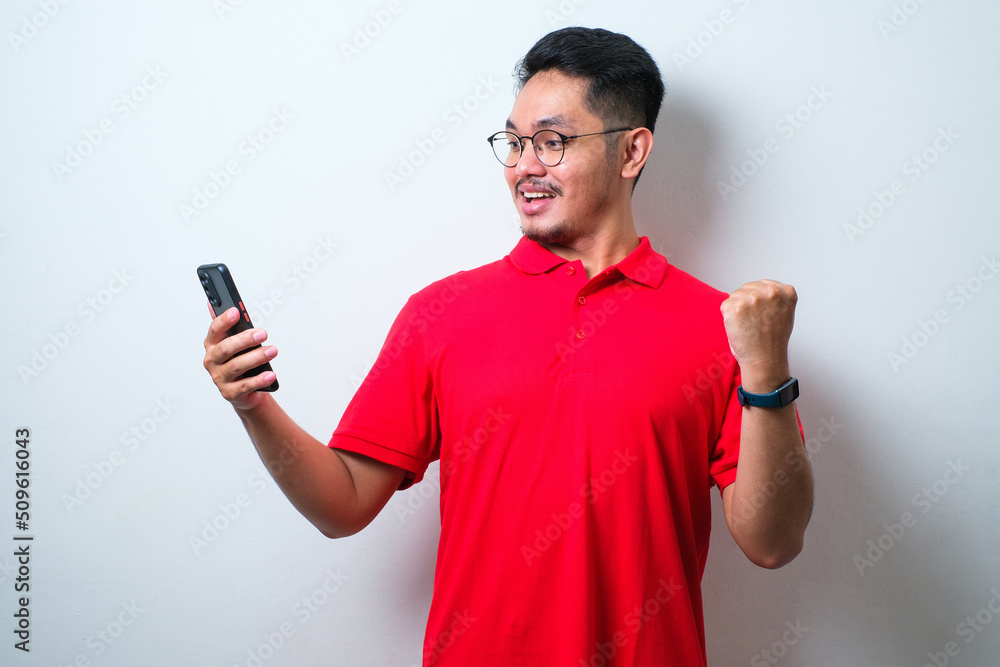 Young handsome man wearing casual shirt and glasses standing very happy and excited doing winner gesture with arms raised, smiliing while looking to his mobile phone Young handsome man wearing casual shirt and glasses standing very happy and excited doing winner gesture with arms raised, smiliing while looking to his mobile phone