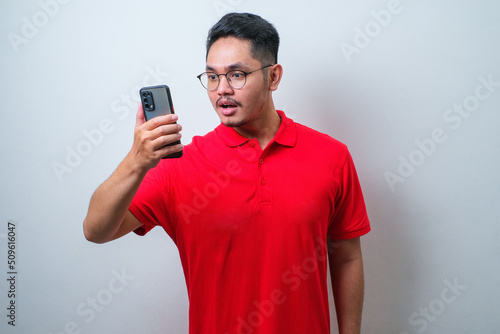 Young handsome Asian man wearing casual shirt and glasses showing shocked face expression while looking to the mobile phone screen