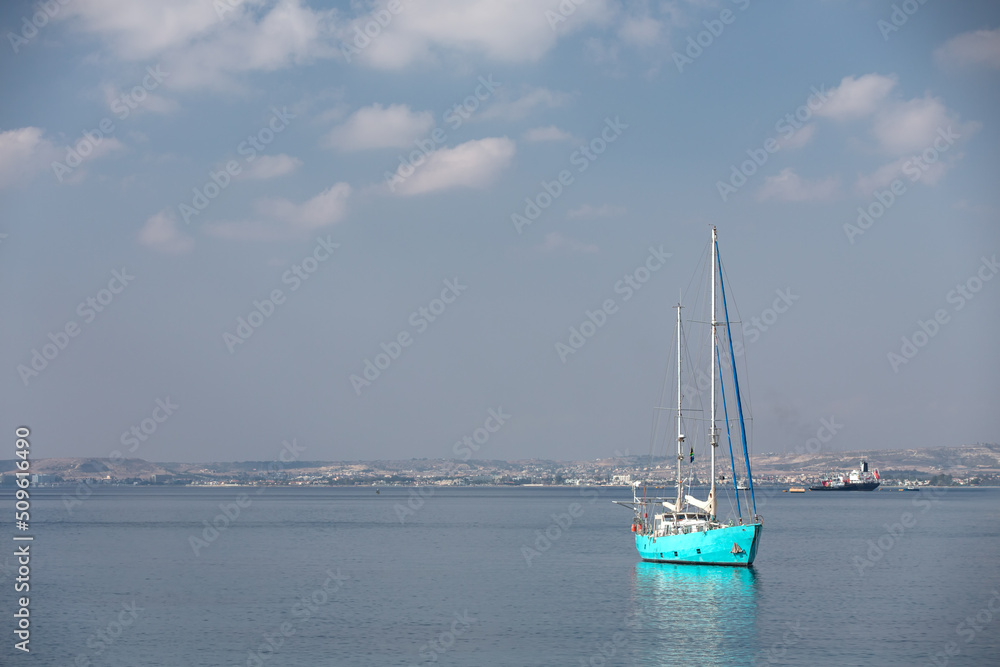A moored boat at sea with lowered sails against the background of the coastline and a cargo ship. Windless weather.