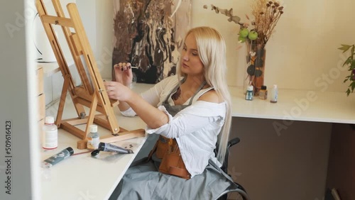 Beautiful girl blonde artist paints an easel while sitting in a wheelchair