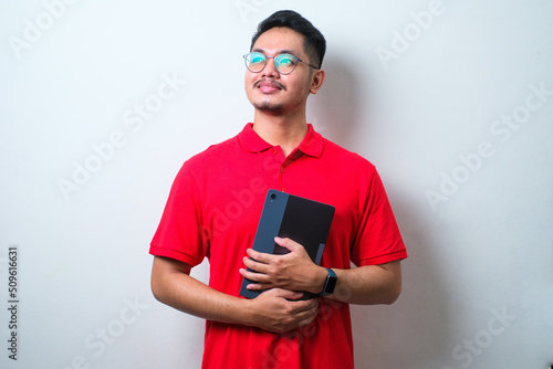 Young handsome Asian man wearing casual shirt and glasses standing, holding tablet looking to the side photo