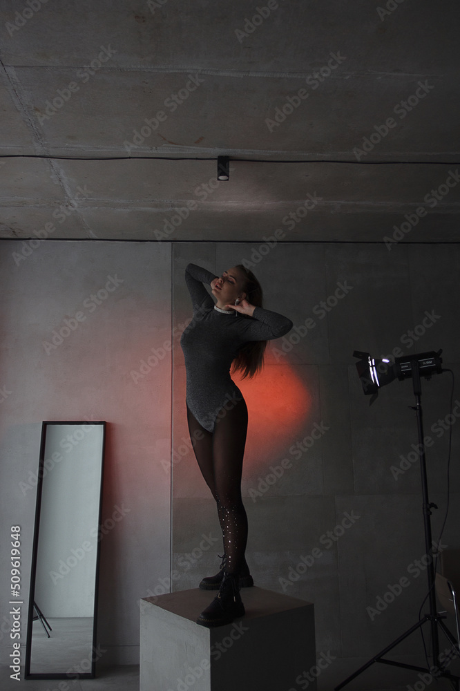 © NASTYA PALEHINA - young teen girl in a shiny silver bodysuit stands fashion on gray cube with hands near head on the gray wall background in loft interior with red studio light and mirror. fashion concept, free space