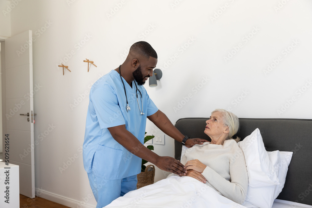 Obraz premium African american male health worker supporting caucasian senior woman lying on the bed at home