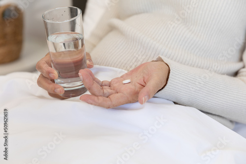 Mid section of caucasian senior woman holding a glass of water and medical pill lying on bed