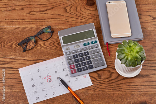 Top view flat lay business accessories on brown wooden desk. Calendar with tax day with calculator and glasses. Green plant in the pot.